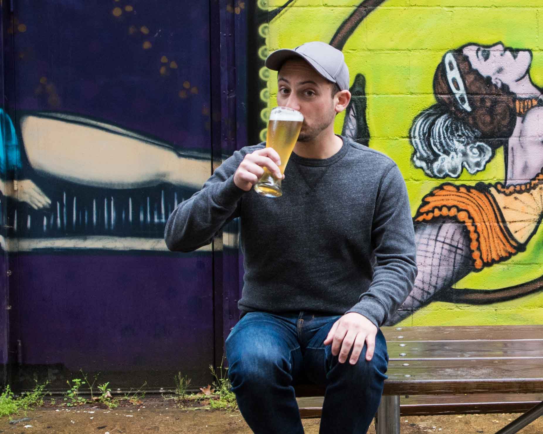 Founder of HiWire Brewing on a picnic table sipping a beer in front of a mural—Mountain BizWorks.