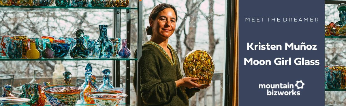 Kristen Muñoz leans against a shelf of her glass work. She is holding a multi-colored glass vase in her hands.