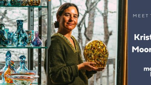 Kristen Muñoz leans against a shelf of her glass work. She is holding a multi-colored glass vase in her hands.